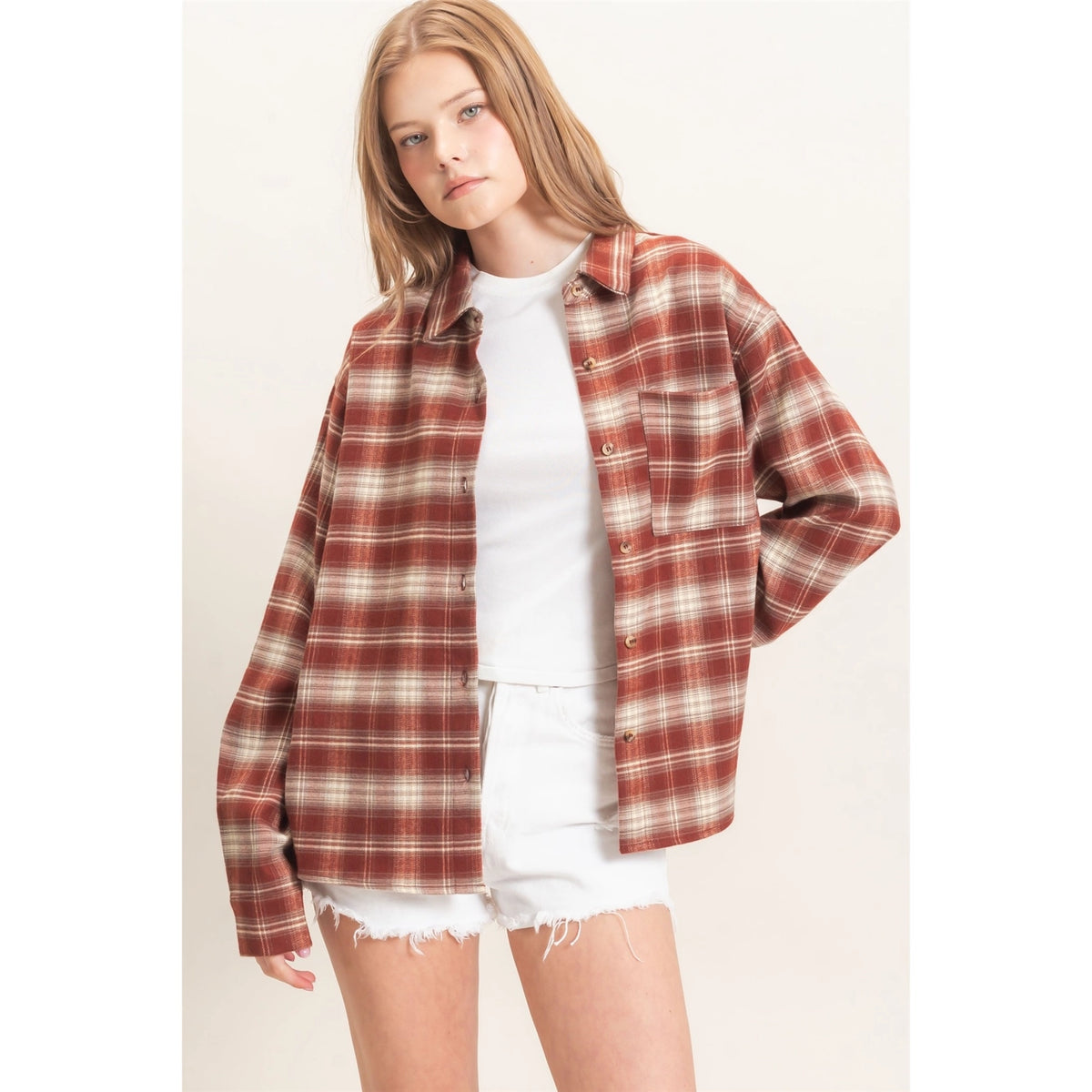A woman modeling an oversized rust red and beige plaid button-up shirt over a white t-shirt and white denim shorts, set against a plain off-white studio background.