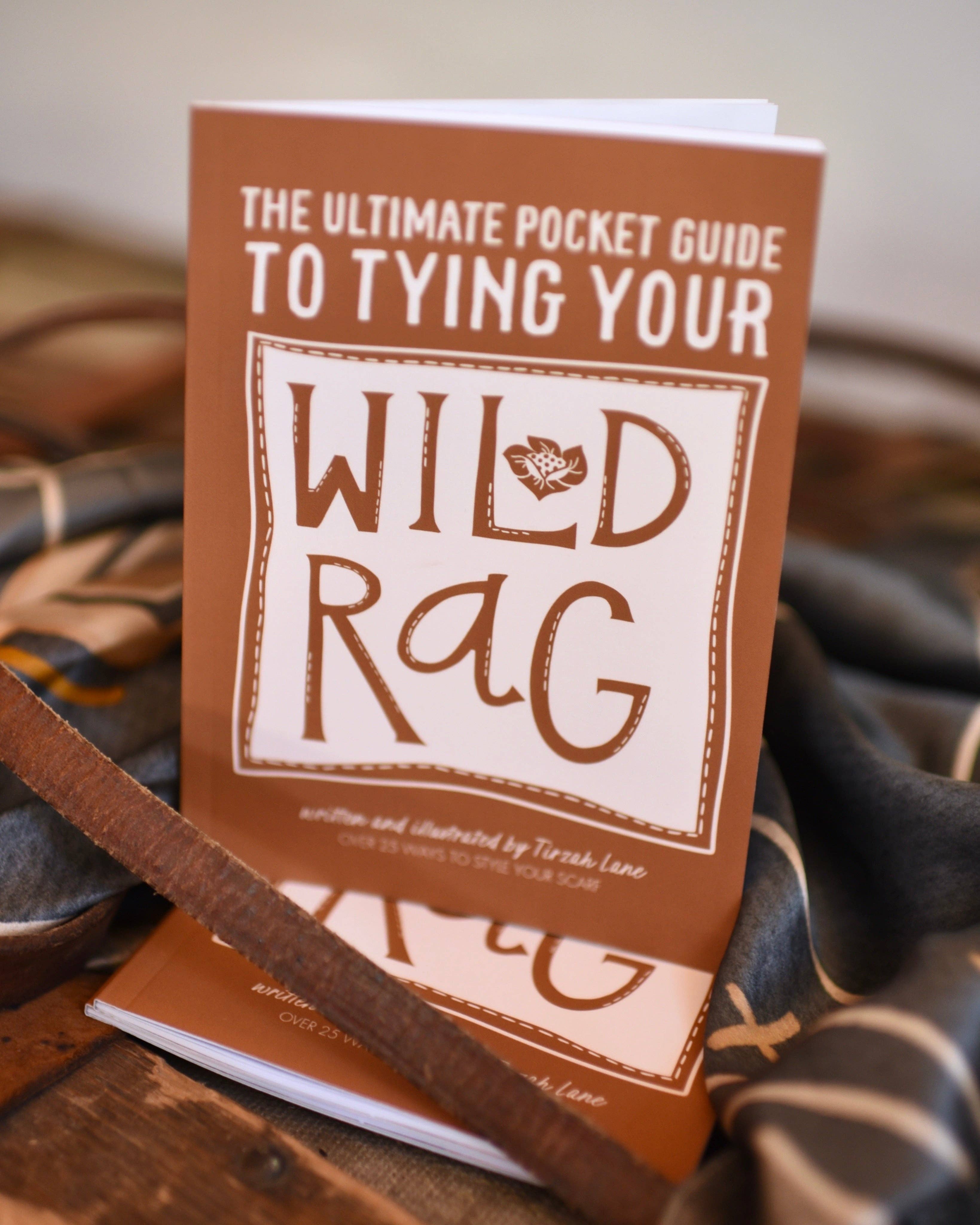 Book titled 'The Ultimate Pocket Guide to Tying Your Wild Rag' on a wooden surface with leather straps.