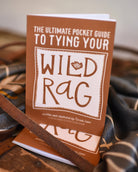 Book titled 'The Ultimate Pocket Guide to Tying Your Wild Rag' on a wooden surface with leather straps.