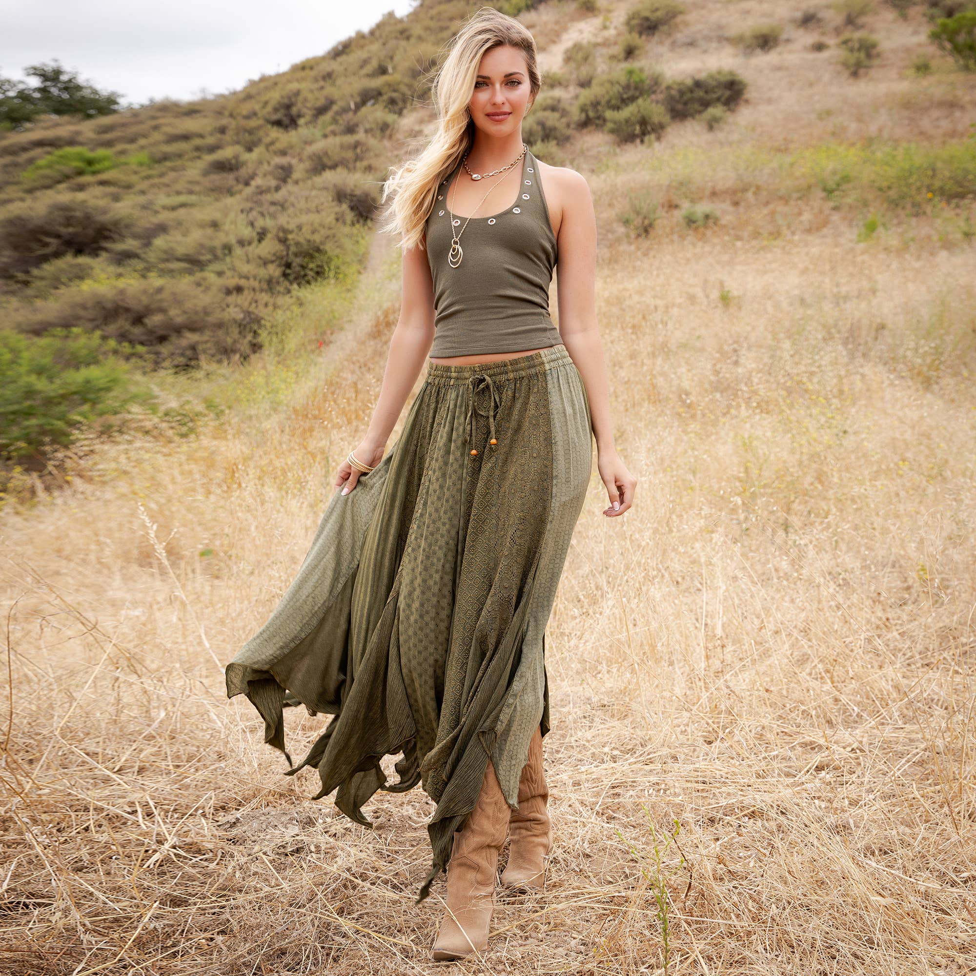 A woman stands in a dry grassy field wearing an olive green halter top and a matching olive green maxi skirt with a drawstring waist, paired with brown boots.