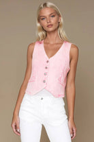 Woman wearing a pink sleeveless top with buttons and white pants on a beige background