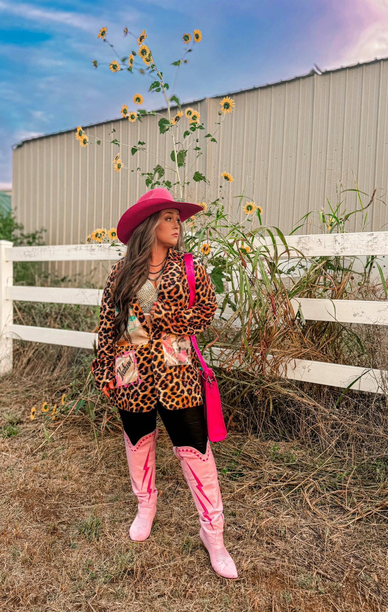 Person wearing a leopard print jacket, pink hat, and boots standing in a sunflower field with a white fence in the background.