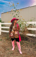 Person wearing a leopard print jacket, pink hat, and boots standing in a sunflower field with a white fence in the background.