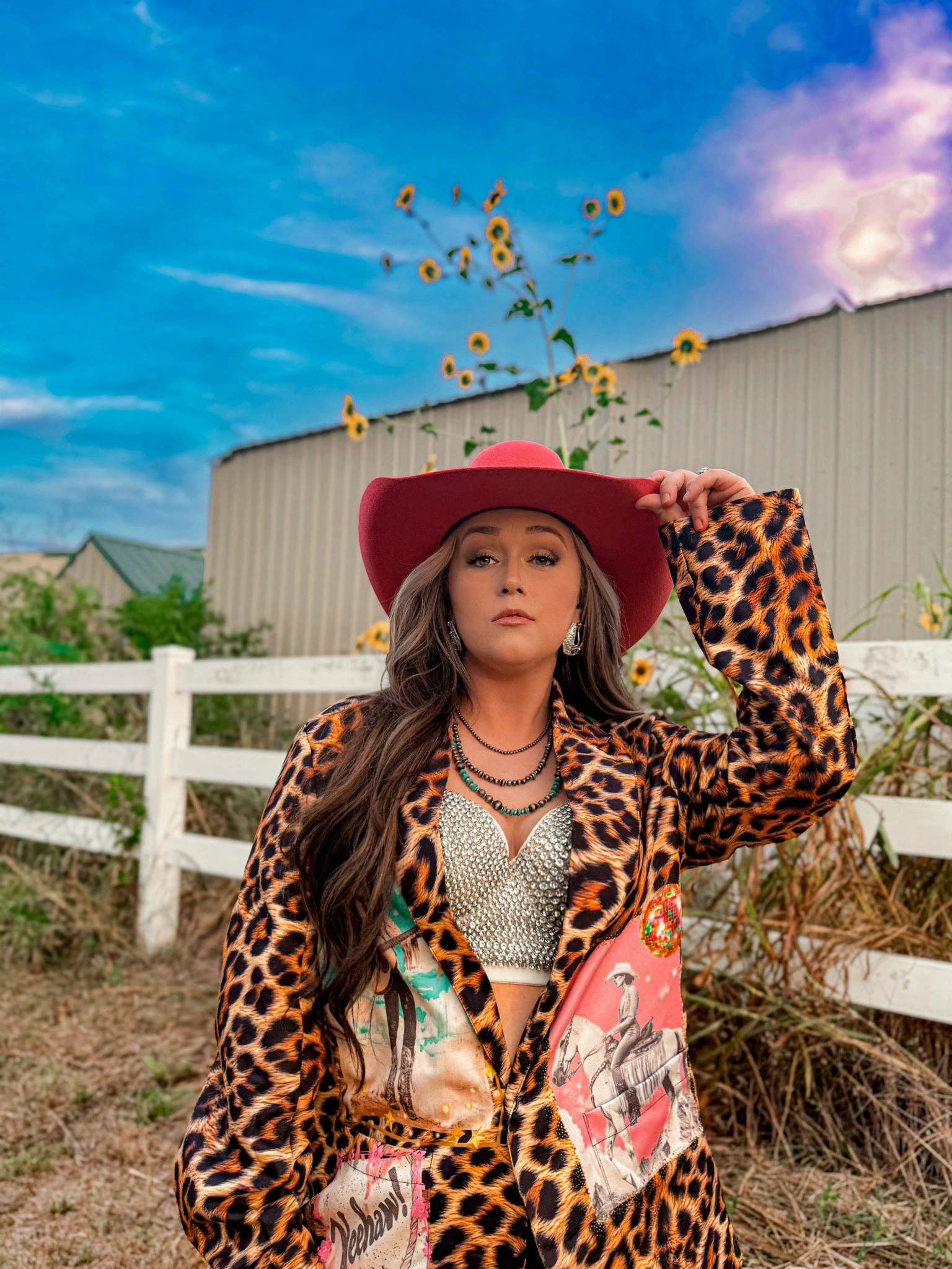 Woman in a leopard print jacket and red hat holding sunflowers against a blue sky.