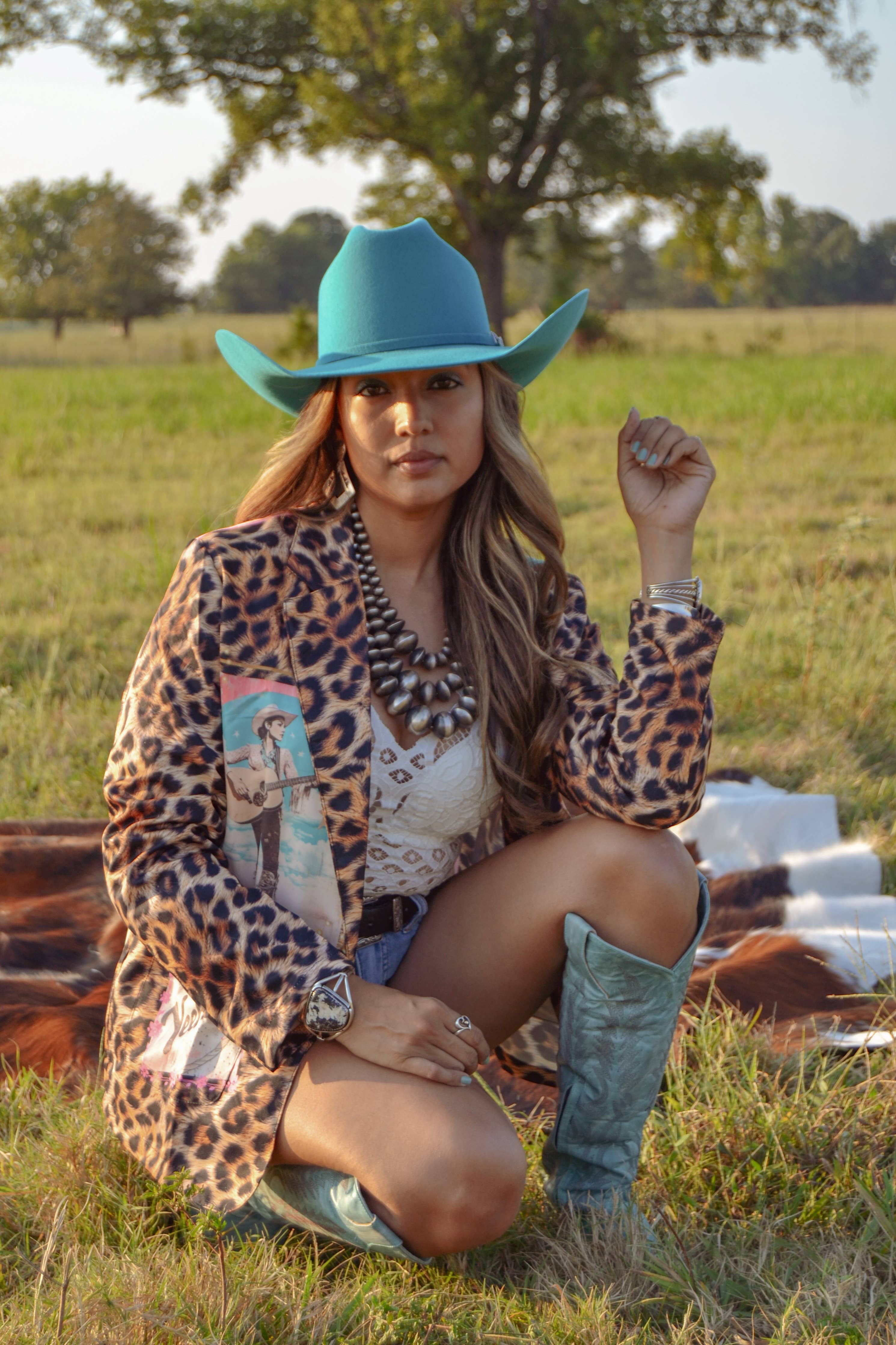 Woman in a leopard print jacket and blue cowboy hat sitting in a field.