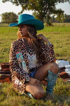 Woman in a leopard print jacket and blue cowboy hat sitting in a field.