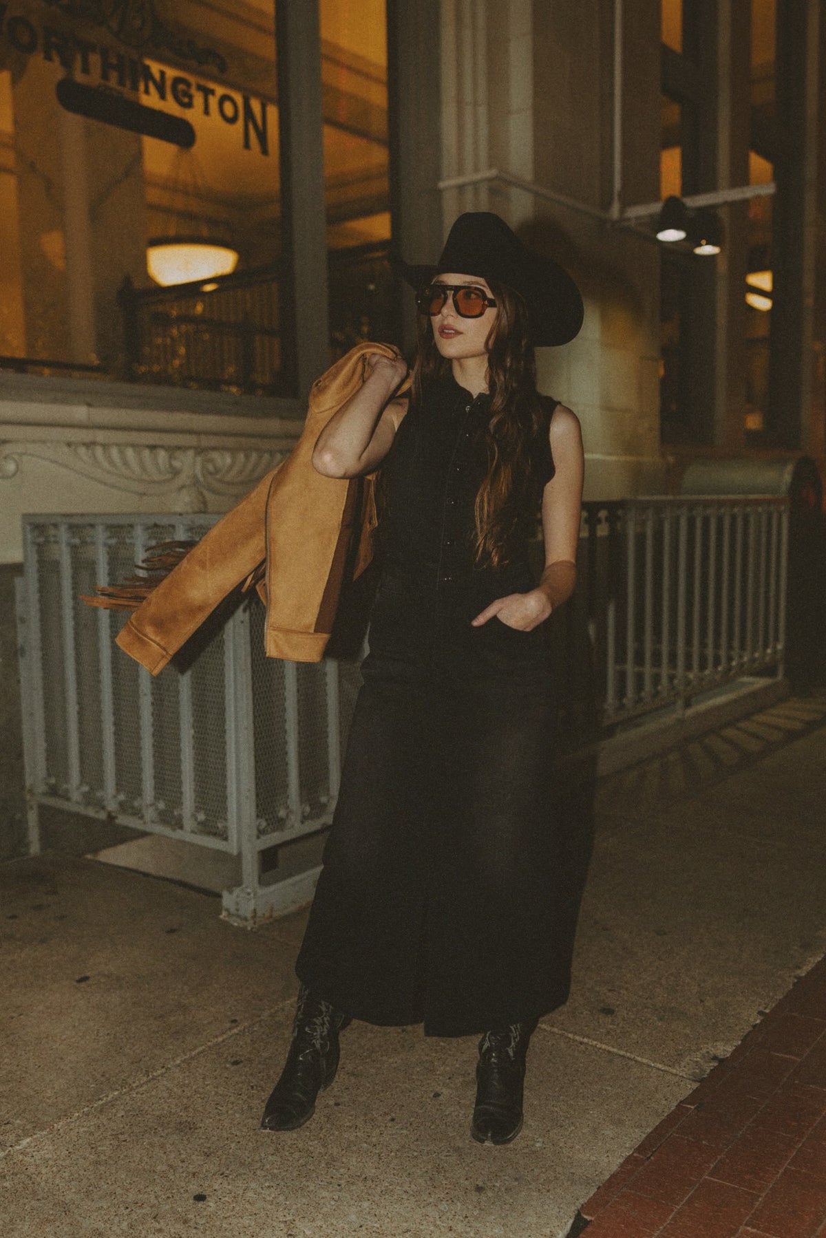 Woman in a black dress and hat standing on a city street at night.