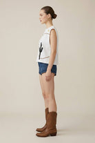 Woman wearing a white sleeveless top with a cactus design, denim shorts, and brown boots on a beige background