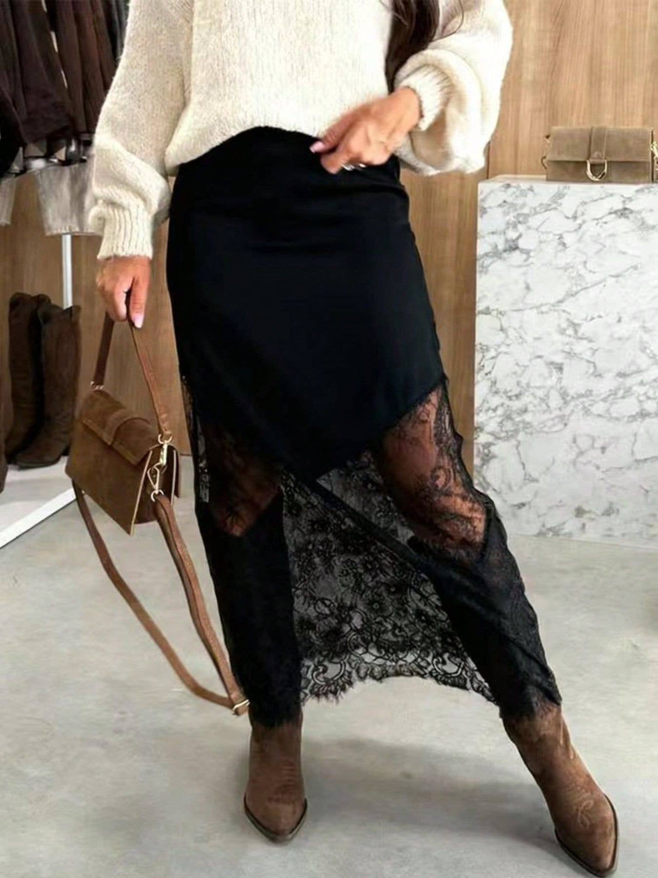 Person wearing a black lace skirt with a white sweater and brown boots, holding a brown handbag.