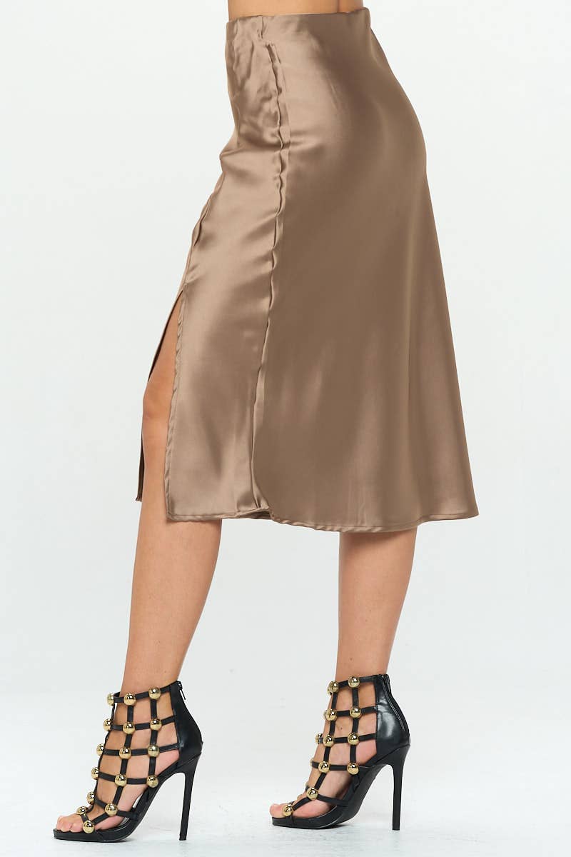 A woman modeling a satin midi skirt with a side slit, in a metallic color, paired with a satin top with a bow detail.