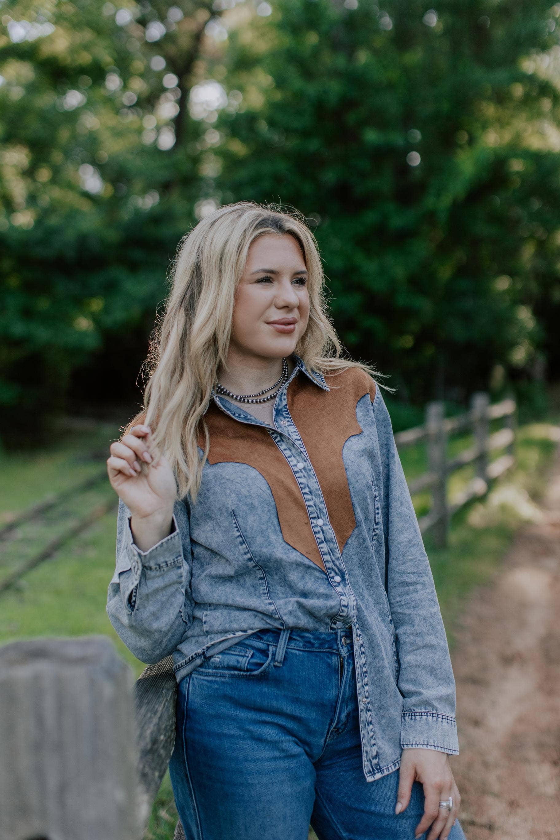 Woman wearing a denim and brown shirt and blue jeans outdoors with greenery in the background