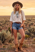 A woman stands in a desert landscape at sunset wearing a wide-brim brown hat, a cream graphic T-shirt, denim shorts, a brown fringe belt, layered turquoise necklaces, and brown cowboy boots.