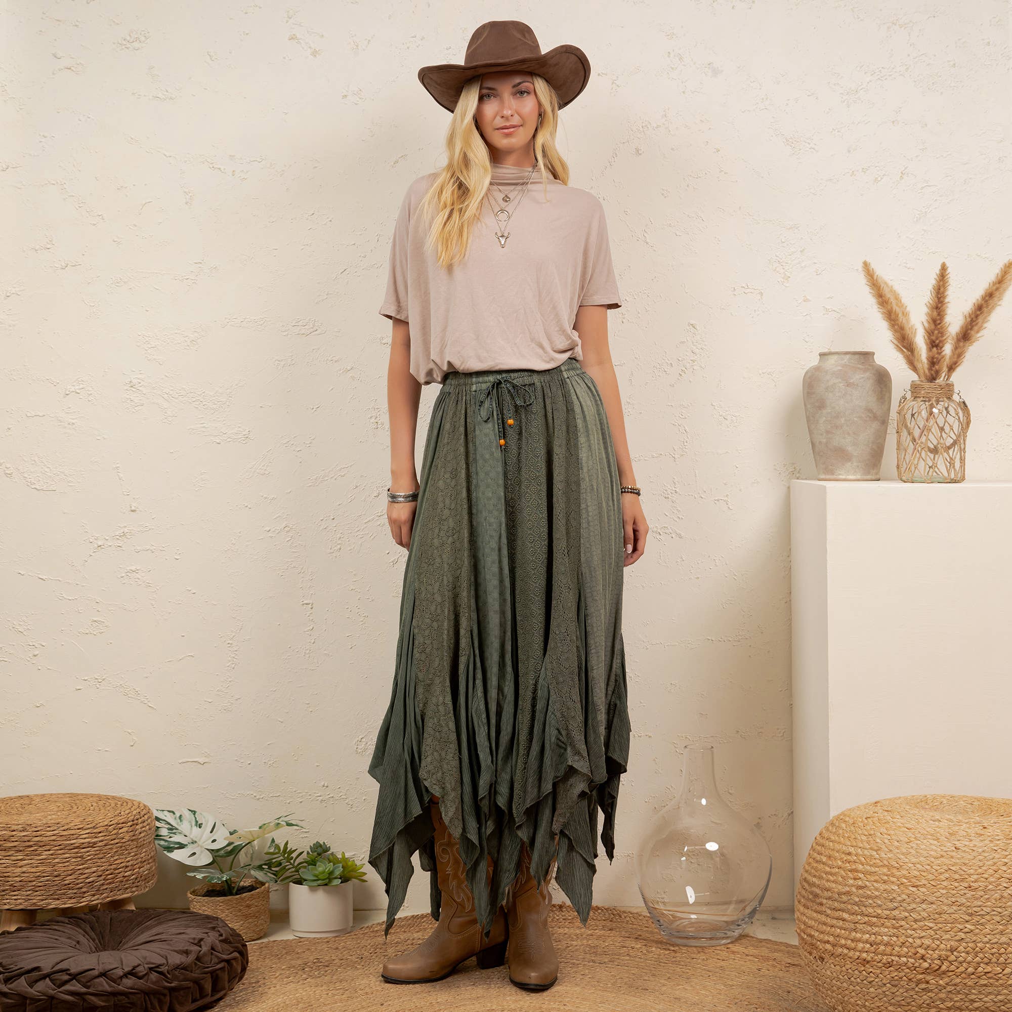 A woman stands indoors wearing a brown cowboy hat, a light beige short-sleeve top, a dark olive green high-low skirt with button details, and brown leather boots. The setting includes a textured cream wall, a white pedestal with dried grass in a vase, and woven poufs.
