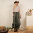 A woman stands indoors wearing a brown cowboy hat, a light beige short-sleeve top, a dark olive green high-low skirt with button details, and brown leather boots. The setting includes a textured cream wall, a white pedestal with dried grass in a vase, and woven poufs.