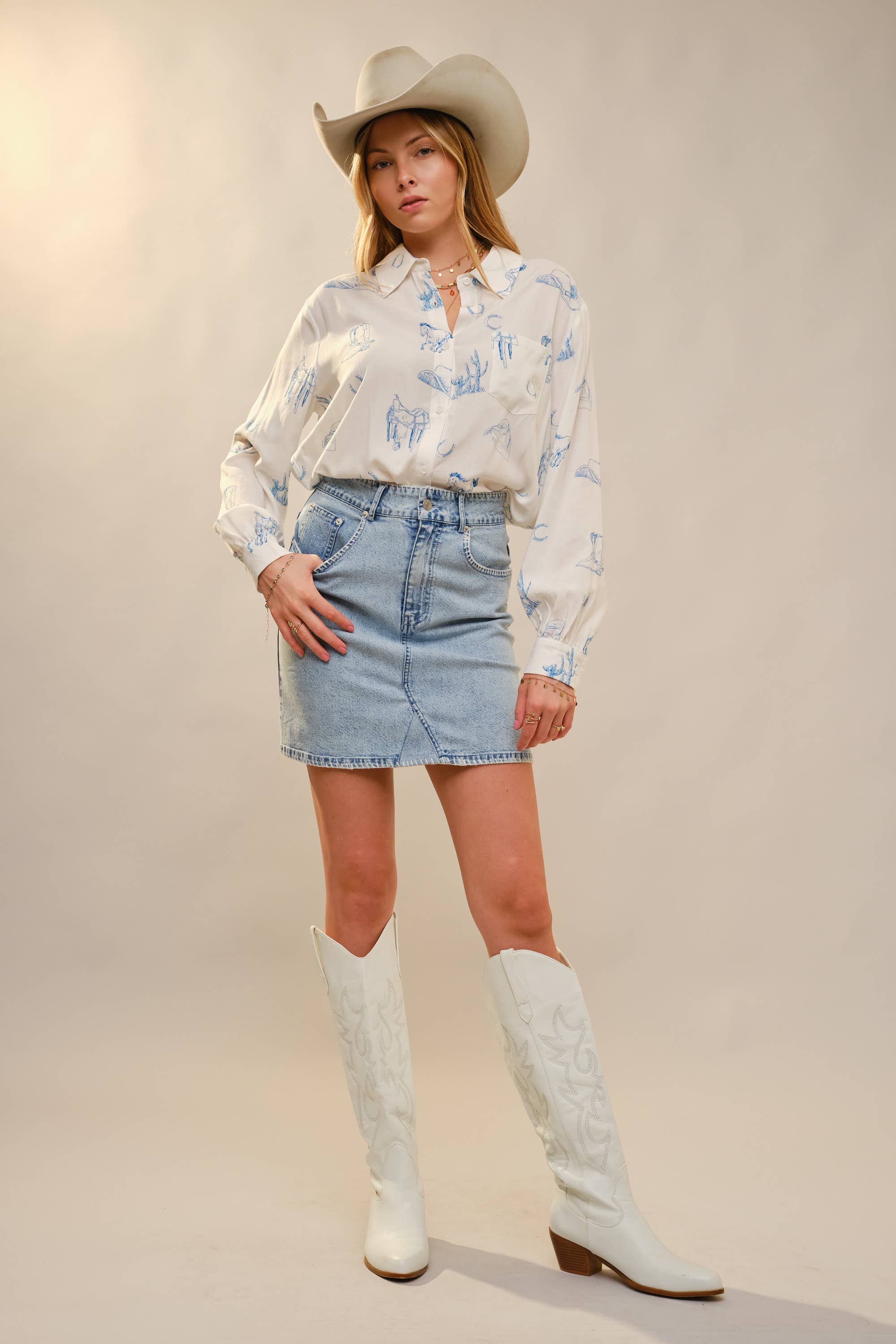 Woman wearing a white shirt with blue patterns, denim skirt, and white cowboy boots against a plain background