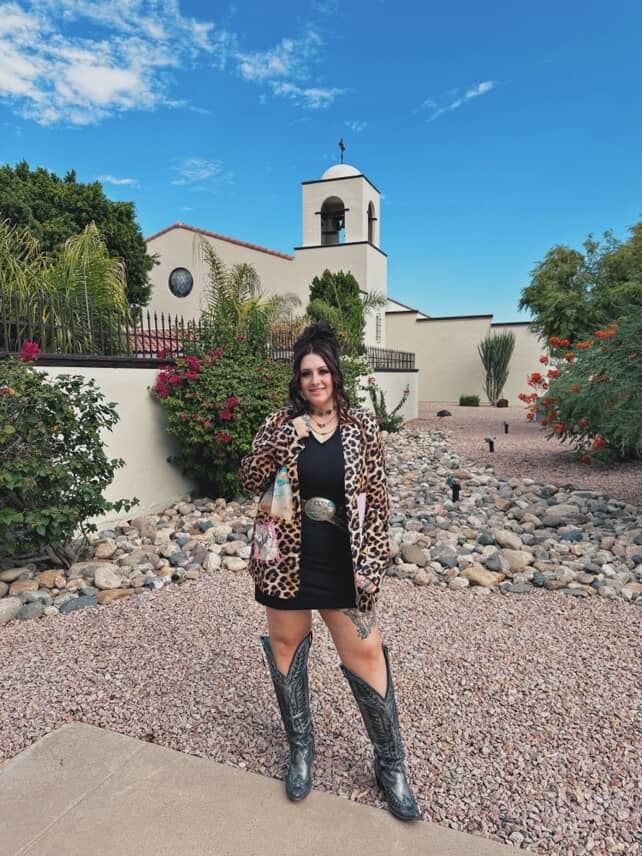Woman in leopard print jacket and black dress standing in front of a church with a bell tower.