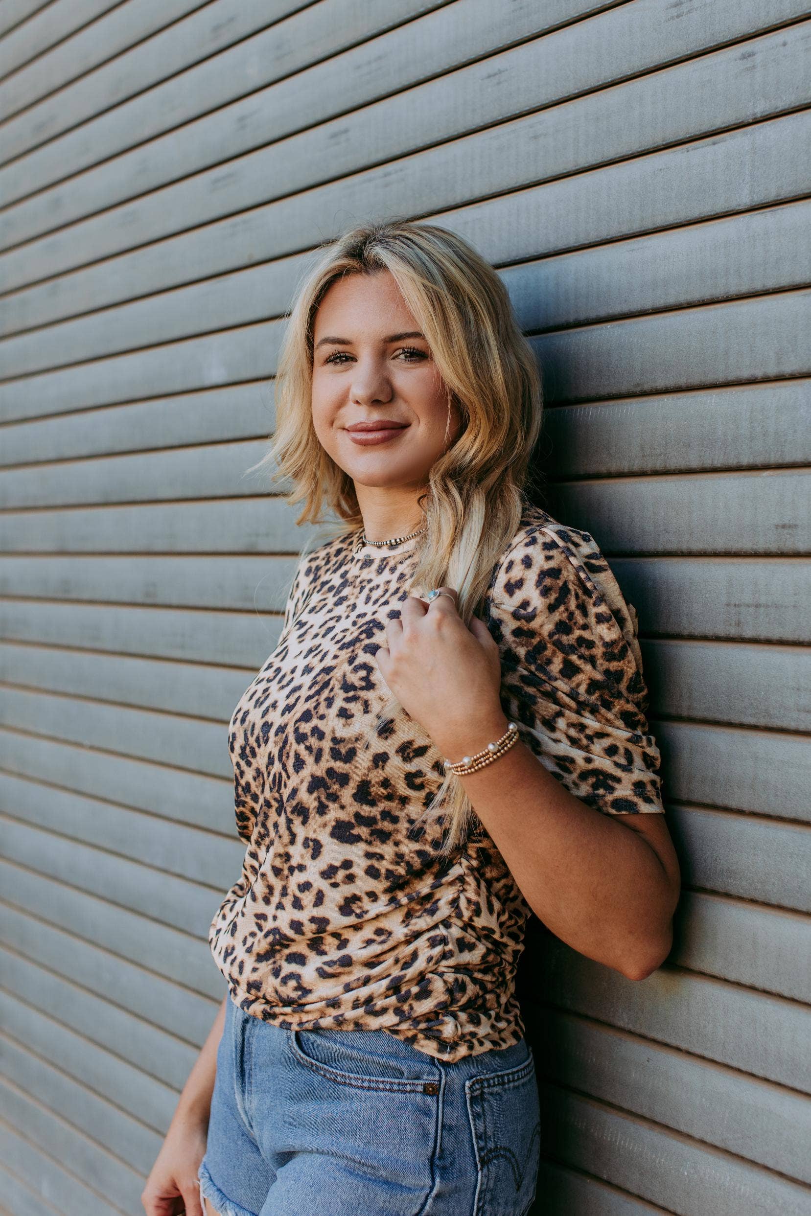 Woman wearing a leopard print top and blue jeans leaning against a wooden wall.