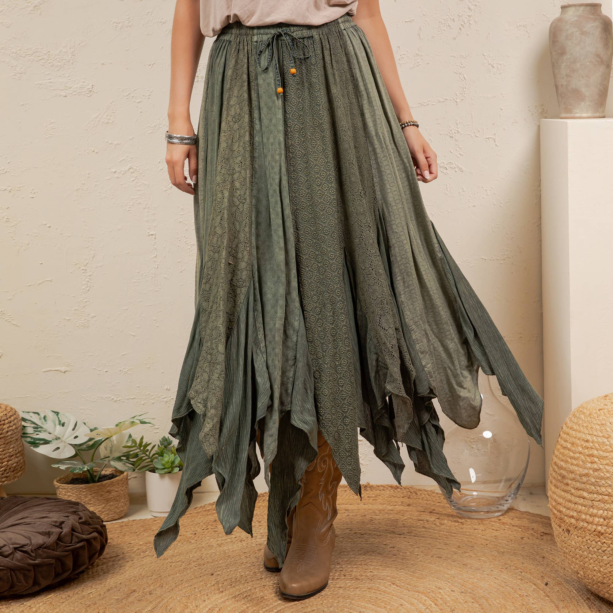 A woman wearing a long, flowing sage green skirt with a tie waist and brown ankle boots, standing in a styled room with plants and woven decor.