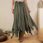 A woman wearing a long, flowing sage green skirt with a tie waist and brown ankle boots, standing in a styled room with plants and woven decor.