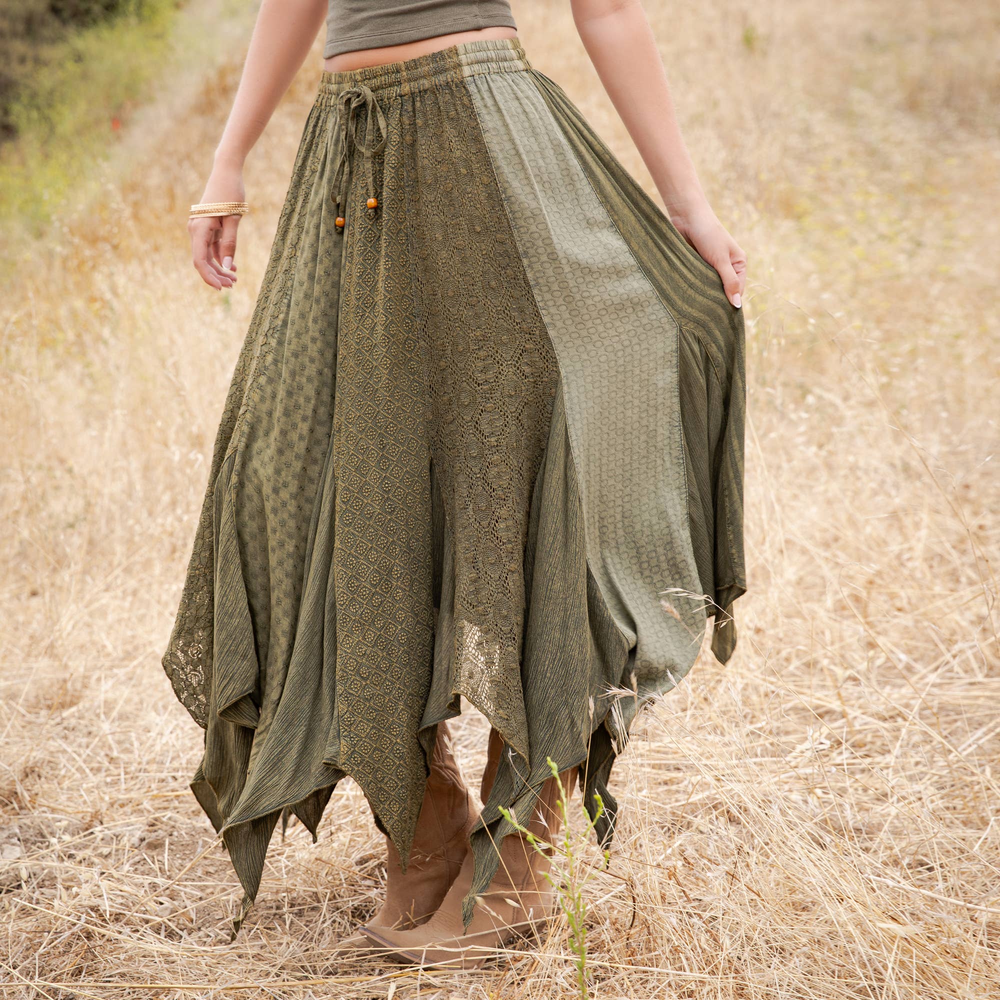 An olive green bohemian maxi skirt with mixed lace and textured fabric panels, worn by a model standing in a dry grass field.