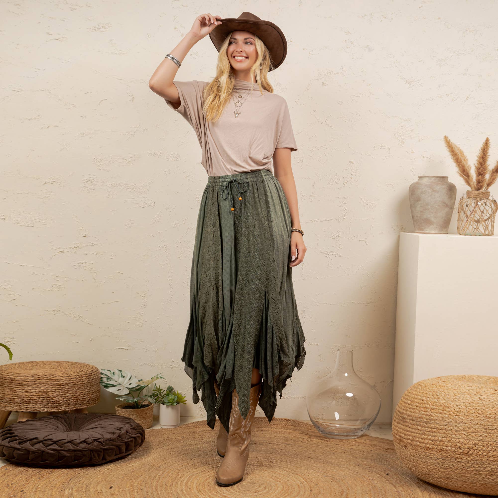 A woman stands in a styled indoor setting wearing a brown wide-brim hat, a light beige short-sleeve top, and a dark olive green high-waisted, tiered maxi skirt with brown leather boots.