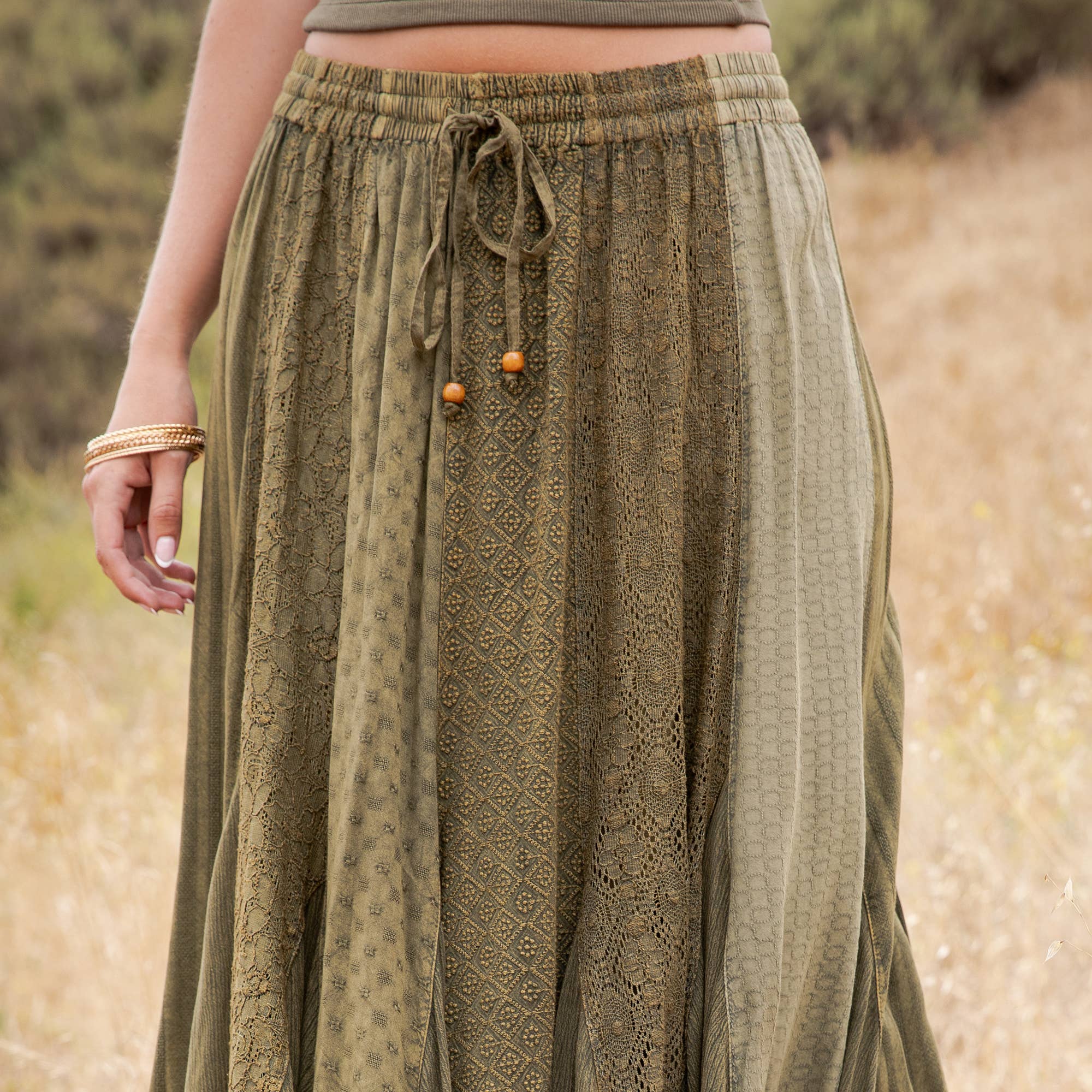 A close-up of a person wearing a loose, olive green patterned skirt with an elastic drawstring waistband, standing outdoors against a blurred natural background.