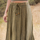 A close-up of a person wearing a loose, olive green patterned skirt with an elastic drawstring waistband, standing outdoors against a blurred natural background.