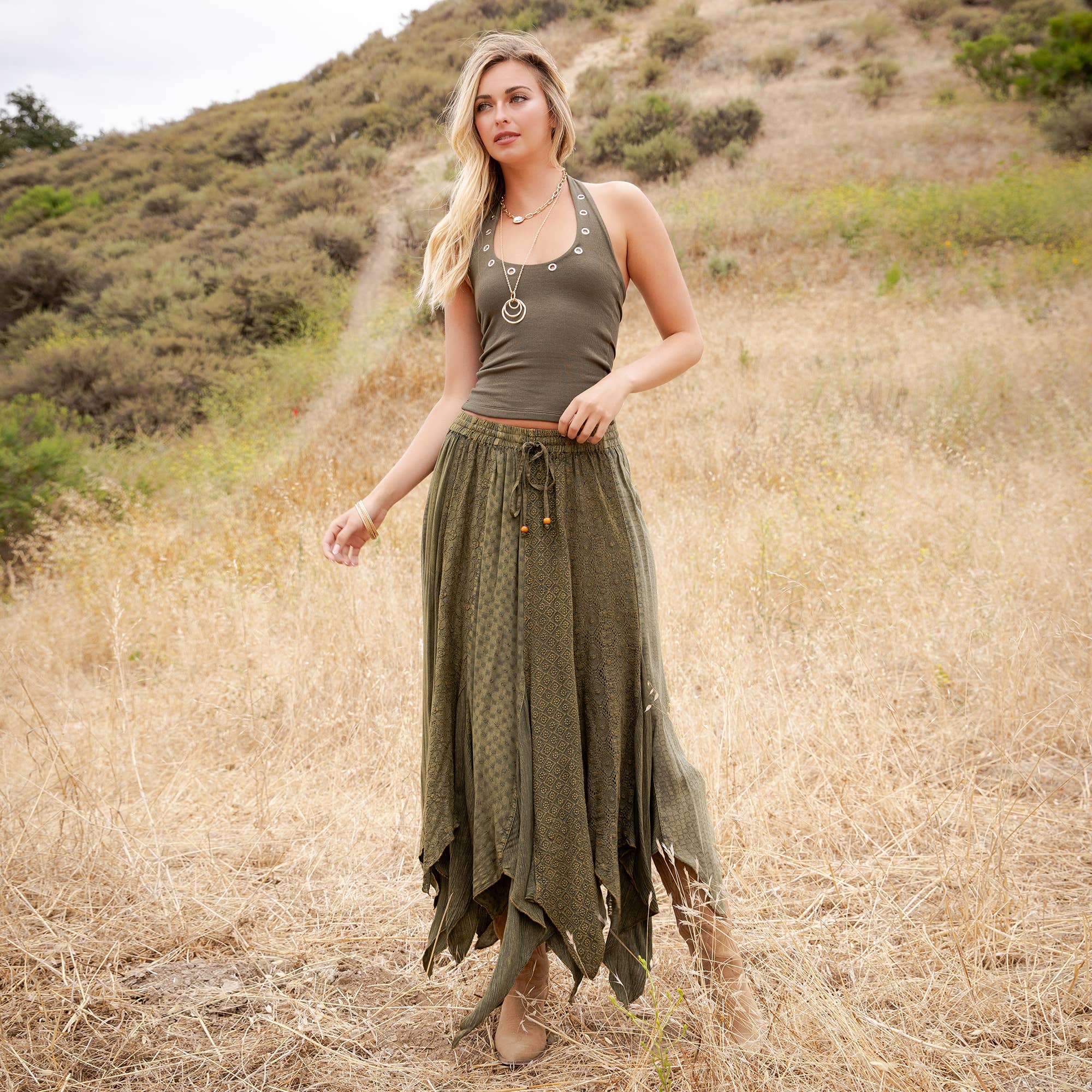 A woman stands in a dry grassy field wearing an olive green tank top and a matching olive green maxi skirt with a tie waist and layered fringe details.