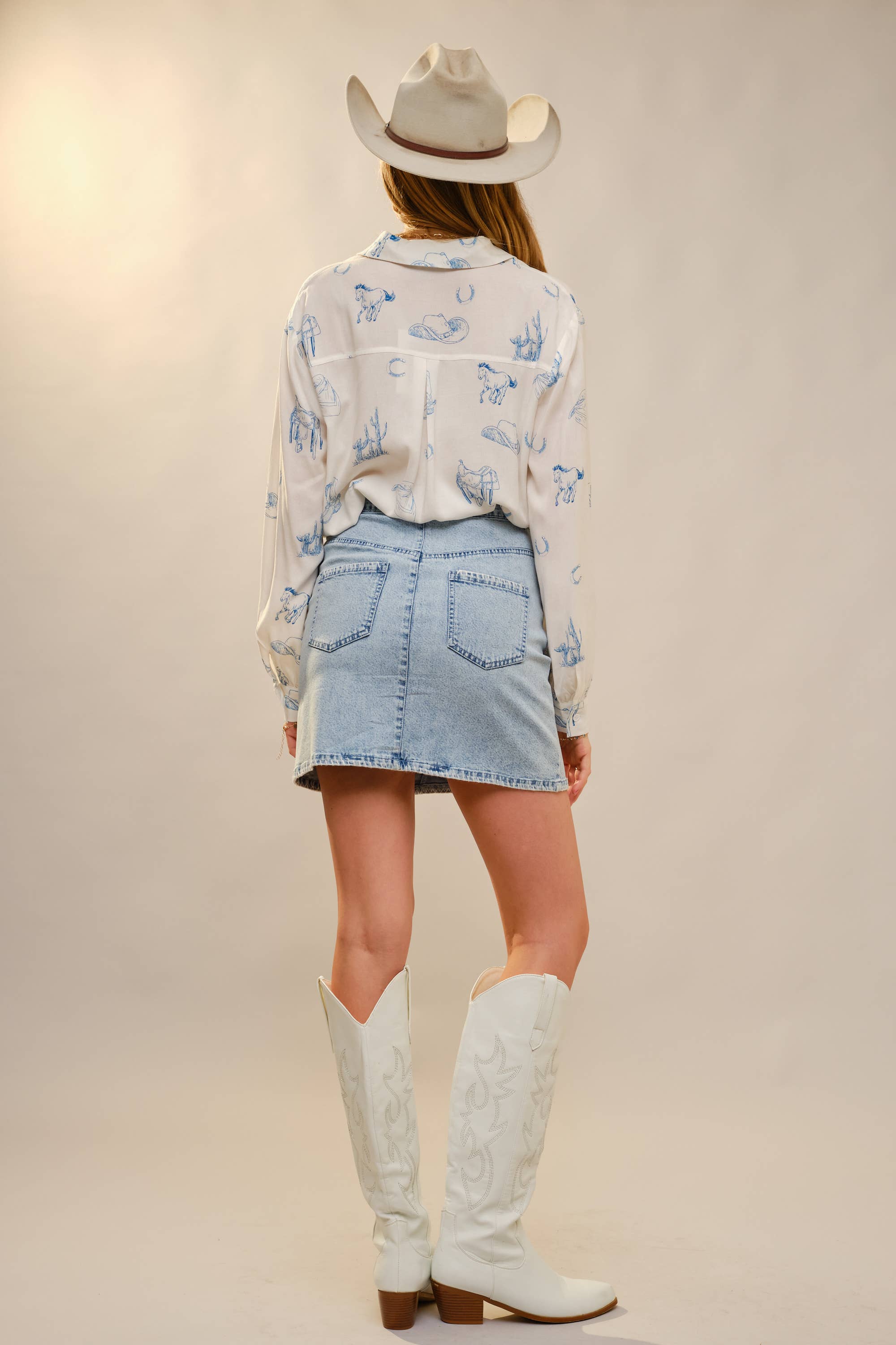 Person wearing a white blouse with blue patterns, denim skirt, and white cowboy boots on a plain background