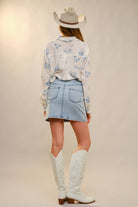Person wearing a white blouse with blue patterns, denim skirt, and white cowboy boots on a plain background