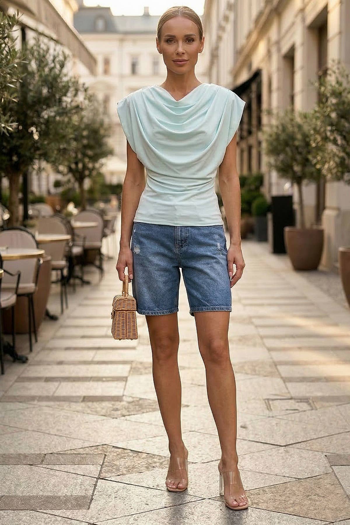 Woman in light blue top and denim shorts standing on a street with outdoor seating in the background