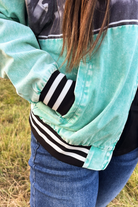 A close-up of a person wearing a mint green and black color-blocked jacket with black and white striped ribbed cuffs and hem, paired with blue jeans, standing outdoors on grass.