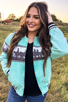 A woman wearing a mint green bomber jacket with a black and white western horse pattern across the chest, standing in an outdoor grassy field at sunset.