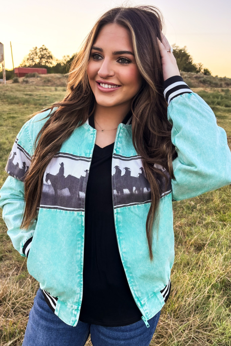 A woman wearing a mint green bomber jacket with a black and white western horse pattern across the chest, standing in an outdoor grassy field at sunset.