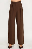 A pair of high-waisted, wide-leg brown trousers shown on a model from the waist down, paired with dark brown pointed-toe heels against a plain light gray studio background.