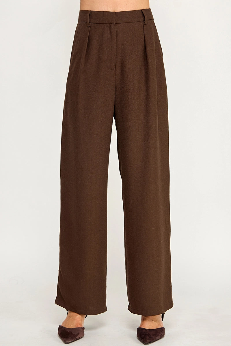 A pair of high-waisted, wide-leg brown trousers shown on a model from the waist down, paired with dark brown pointed-toe heels against a plain light gray studio background.