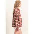 A woman shown in side profile wearing an oversized long-sleeve plaid button-up shirt in rust red, beige, and white, paired with white frayed denim shorts against a plain off-white studio background.