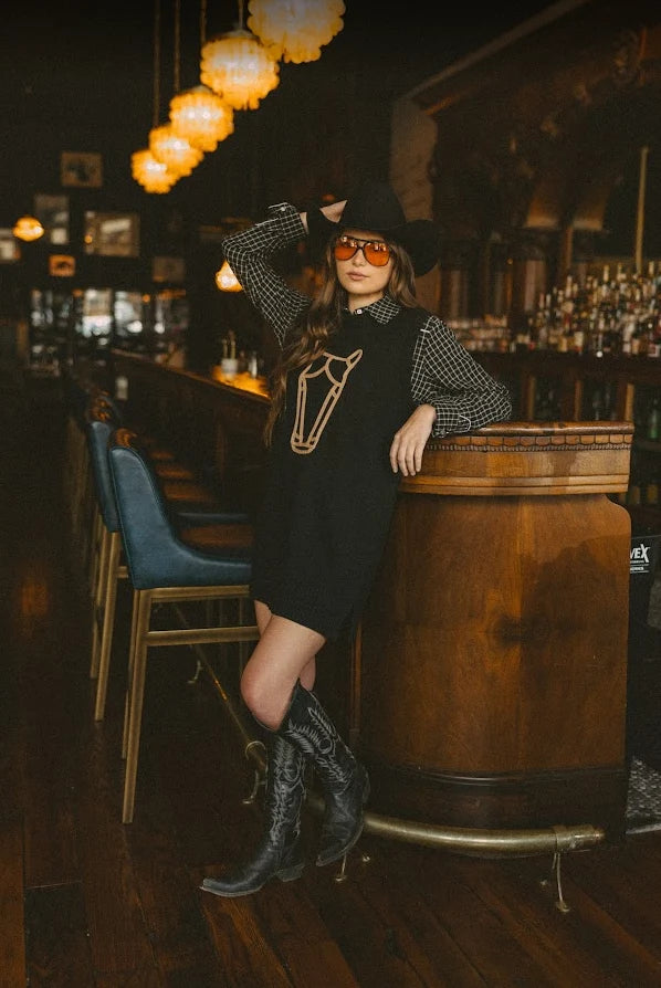 A woman posing in a rustic bar setting wearing a black cowgirl-style dress with a horse graphic, a black-and-white checkered long-sleeve shirt, a black cowboy hat, and black cowboy boots.