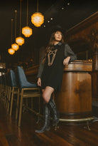 A woman wearing a black cowboy-style mini dress with a gold horse outline, a black-and-white gingham shirt, a black cowboy hat, and black leather boots, leaning against a wooden bar in a dimly lit bar setting.