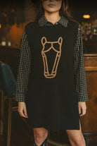 A woman wearing a black knit sweater vest over a black-and-white checkered long-sleeve shirt, with a tan embroidered horse head outline on the front, photographed in a dimly lit indoor setting.
